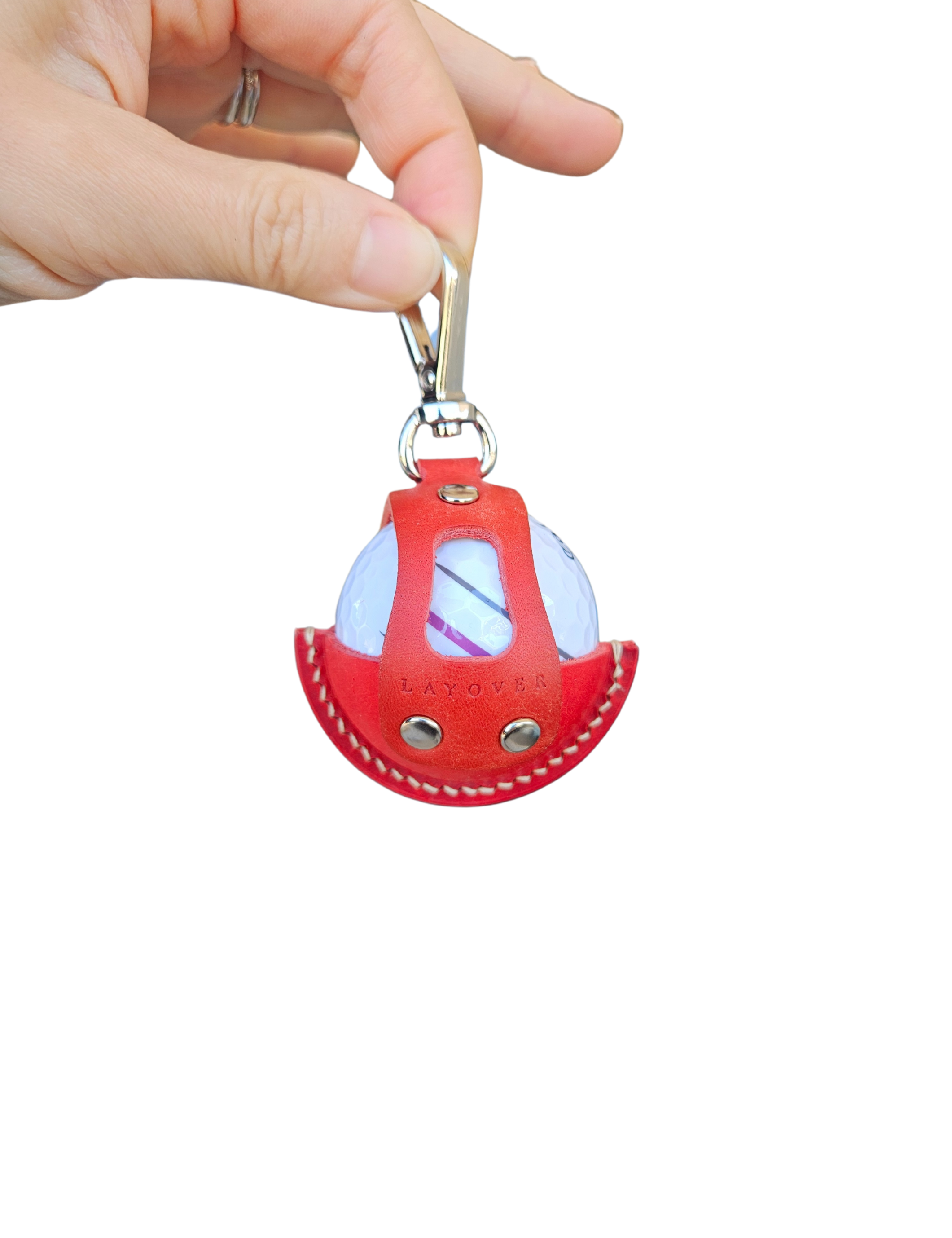 A close-up of a red leather golf ball holder with visible stitching and metallic rivets, held by a hand. The holder features a secure clip at the top and an open-front design showcasing a white golf ball inside